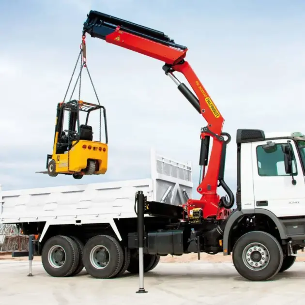 employé de chantier qui est en train de ranger un chariot élévateur dans un camion aidé du grues auxiliaires de chargement